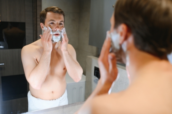 A shirtless man wrapped in a towel applies shaving cream to his face while looking in the mirror, mid-grooming routine in a modern bathroom setting.