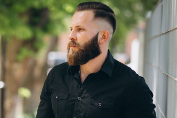 Bearded man with a well-groomed, full beard and neatly styled hair stands outdoors wearing a black button-up shirt, exuding confidence and showcasing even beard growth.