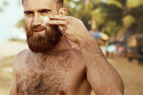 Bearded man on a sunny beach applying sunscreen to his face, highlighting skin and beard protection under harsh UV rays. He stands shirtless with palm trees and blurred beachgoers in the background, capturing a relaxed, tropical setting.