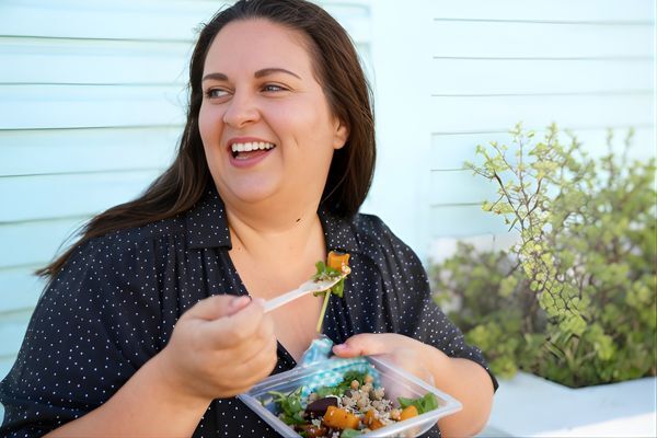 A woman with long brown hair smiles while enjoying a fresh salad outdoors, wearing a navy polka-dot blouse. She appears relaxed and happy, embracing a balanced lifestyle—an image that aligns with Hair Mayraki’s holistic approach to beauty and wellness.