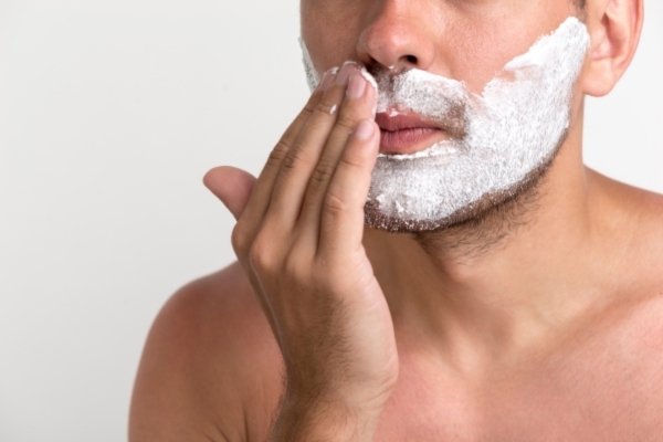 A close-up of a shirtless man applying thick white shaving cream to his upper lip and cheeks, highlighting a grooming moment in progress.