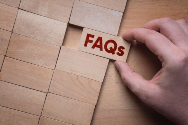A hand placing a wooden block labeled “FAQs” in red letters into a wooden grid, representing a frequently asked questions section or help guide.