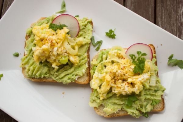 Two slices of toast topped with mashed avocado, scrambled eggs, radish slices, and fresh herbs are served on a white plate. The dish is presented on a rustic wooden table, showcasing a nutritious meal rich in healthy fats and protein.