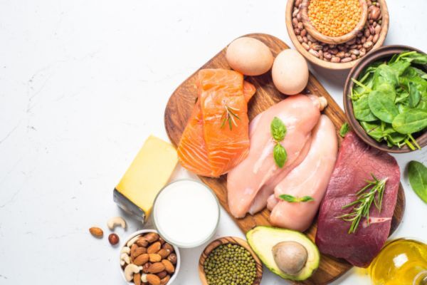 A selection of high-protein foods including raw salmon, chicken breast, beef, eggs, avocado, cheese, milk, nuts, seeds, lentils, and spinach arranged on a wooden board and white surface.