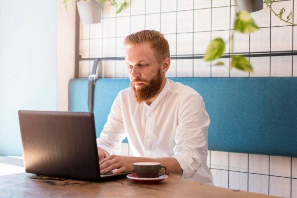 A man with a red beard and white shirt works on a laptop in a modern café setting with a cup of coffee in front of him. He’s seated in a bright space with a grid wall and hanging plants in the background.