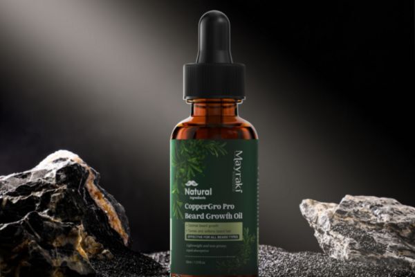 A bottle of Mayraki Natural CopperGro Pro Beard Growth Oil stands between dark mineral rocks under a spotlight, highlighting its earthy, premium branding.