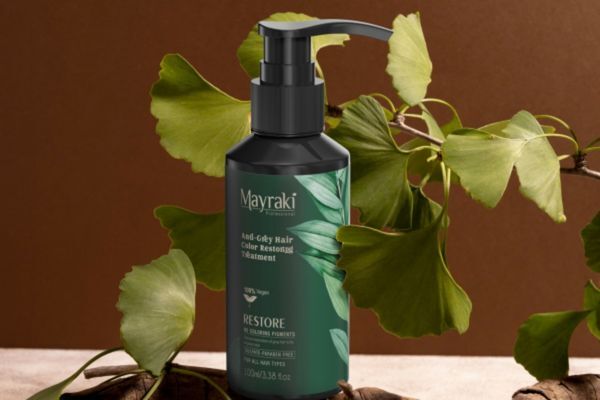Going Back to Your Roots A bottle of Mayraki Anti-Grey Hair Color Restoring Treatment stands upright against a warm brown background, surrounded by fresh green ginkgo biloba leaves. The sleek dark green bottle features botanical graphics and product details in white text, highlighting it as a vegan formula designed to restore natural hair color and prevent greying.