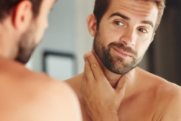 A man with a trimmed beard looks at himself in the mirror while gently touching his neck and jawline, appearing pleased with his skin’s condition. The scene suggests a grooming or skincare routine focused on achieving healthier skin.