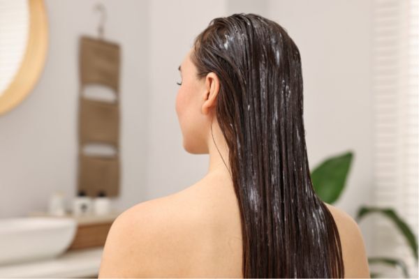 A woman with her back to the camera sits in a bathroom with visible hair treatment cream applied to her long, wet hair, illustrating a deep conditioning or moisturizing hair care routine.