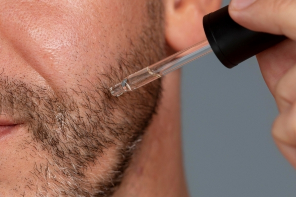 Close-up of a man's face as he applies beard oil using a dropper along the edge of his trimmed beard.