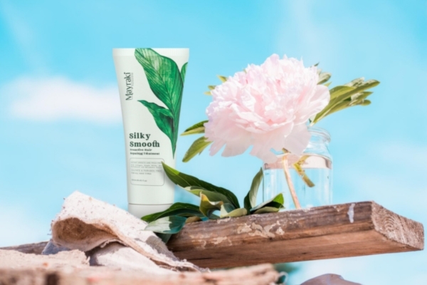 A tube of Mayraki Silky Smooth Proactive Hair Repair Treatment stands on a rustic wooden shelf beside a glass jar holding a soft pink peony. Set against a bright blue sky, the scene evokes freshness and natural care, highlighting the product's focus on restoring damaged hair with gentle, plant-based ingredients.