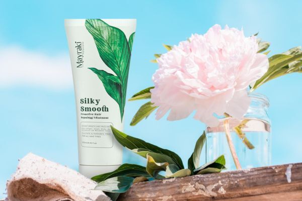 A tube of Mayraki Silky Smooth Proactive Hair Repairing Treatment stands upright beside a pink peony in a glass jar, set against a bright blue sky. The product packaging features green botanical artwork, emphasizing a natural, nourishing hair care formula.