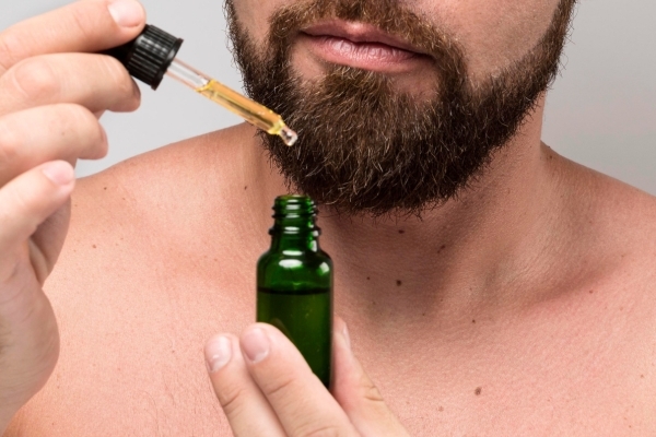 How Natural Oils Prevent and Repair Beard Breakage A close-up of a man applying beard oil from a dropper bottle to his thick, well-groomed beard, illustrating how natural oils prevent and repair beard breakage by hydrating and strengthening the hair.