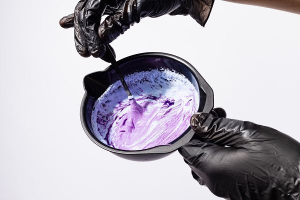 Gloved hands mixing a vibrant purple and white hair dye or toner in a black bowl using a small applicator brush. The creamy mixture suggests preparation for coloring or lightening hair, often part of a salon or at-home dye process.