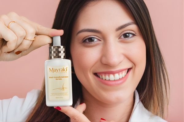 A smiling woman holds up a bottle of Mayraki Hair Growth Intense Serum, pointing to it confidently against a soft pink background. The product presentation supports the role of vitamins and supplements in hair growth through topical applications.