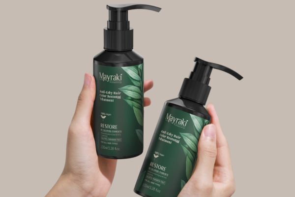 Two hands holding Mayraki Anti-Grey Hair Color Restoring Treatment bottles, featuring dark green labels with leafy designs and black pump dispensers, shown against a neutral beige background.