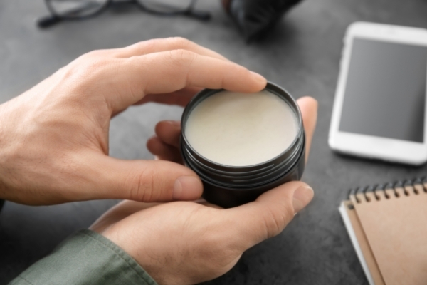 Close-up of a person holding a small black jar of balm, with a creamy product inside. A smartphone, notebook, and glasses are visible in the background on a desk.