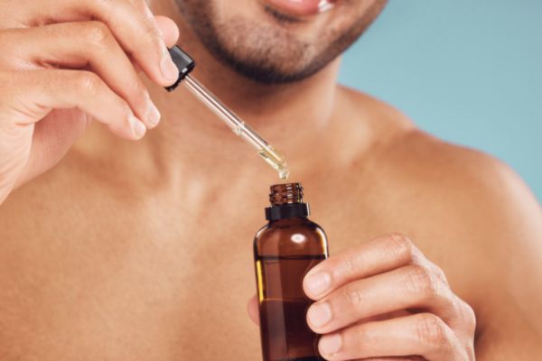 A shirtless man smiles slightly as he uses a dropper to dispense oil from a small amber bottle. The close-up captures a moment of self-care, likely part of a grooming or skincare routine.