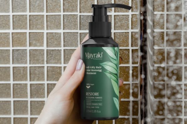 A smiling woman with long, dark hair holds up two Mayraki hair care products—“Ultra Repairing” and “Silky Smooth Frizz-Free Hair Treatment Shampoo”—in each hand. The white and green packaging features botanical designs, emphasizing a natural approach to hair repair and smoothing.