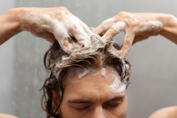 A man with closed eyes massages shampoo into his scalp with both hands, creating a thick lather in his wet hair. The image conveys a sense of cleanliness and scalp care, possibly tied to exfoliation or deep cleansing.