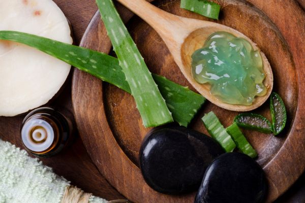 A wooden spoon filled with aloe vera gel rests on a wooden tray alongside fresh aloe vera slices, black massage stones, a small amber dropper bottle, and a soap bar. The setup suggests a natural skincare or beard care routine using aloe vera.