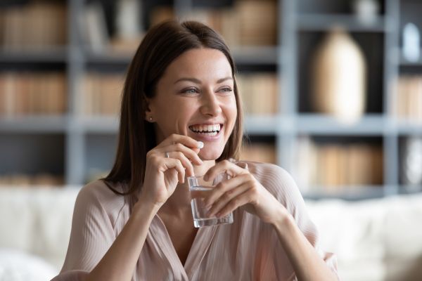 A smiling woman with straight brown hair sits in a cozy room with bookshelves in the background, holding a glass of water and a collagen supplement capsule. She appears relaxed and happy, mid-laugh, as she prepares to take the supplement.