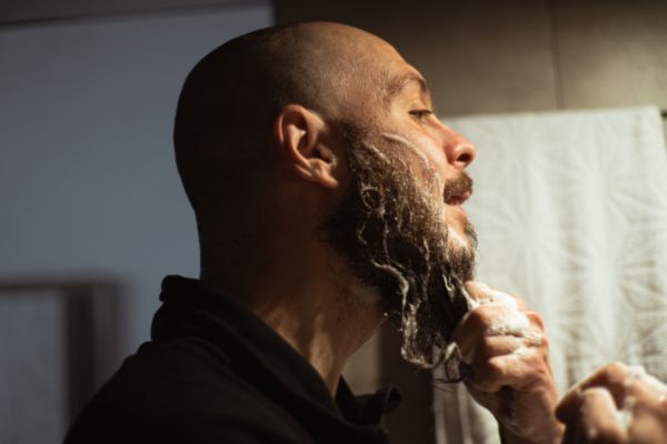 A bald man with a full beard lathers his facial hair with cleansing foam while standing in front of a bathroom mirror, demonstrating beard grooming and care using leave-in conditioner.