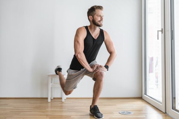 A fit man with a short beard performs a single-leg squat or lunge exercise indoors, balancing with one leg extended behind him. He wears a black tank top and gray shorts, focusing on maintaining his form in a bright, minimalist room with wooden floors and natural light.