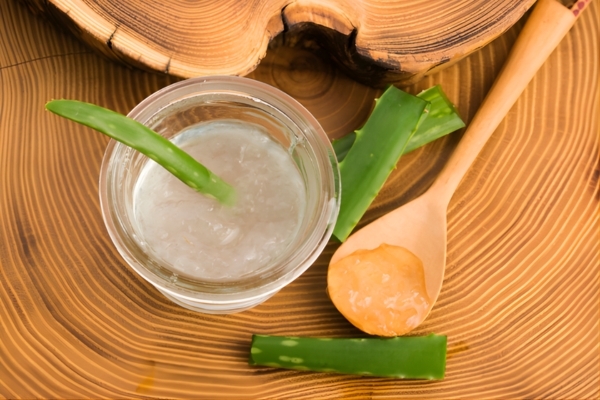 How to Use Aloe Vera for an Itchy Scalp? A glass bowl filled with fresh aloe vera gel sits on a wooden surface, surrounded by sliced aloe leaves and a wooden spoon holding a scoop of the gel. The natural setting emphasizes aloe vera's soothing properties, making it ideal for calming an itchy scalp.