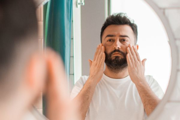A man with a trimmed beard applies skincare product to his cheeks while looking at himself in the mirror, wearing a white t-shirt in a bright bathroom.