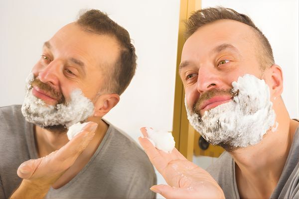 A man stands in front of a mirror, smiling as he applies a thick layer of shaving foam to his beard. This grooming step pairs well with using beard oil for beard growth to maintain softness and skin health post-shave.