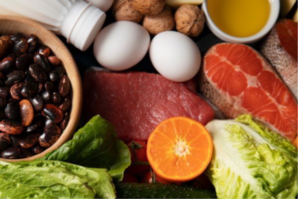 A colorful spread of protein- and vitamin-rich foods including raw salmon, beef, black beans, eggs, lettuce, orange slices, walnuts, and olive oil, suggesting a balanced diet.
