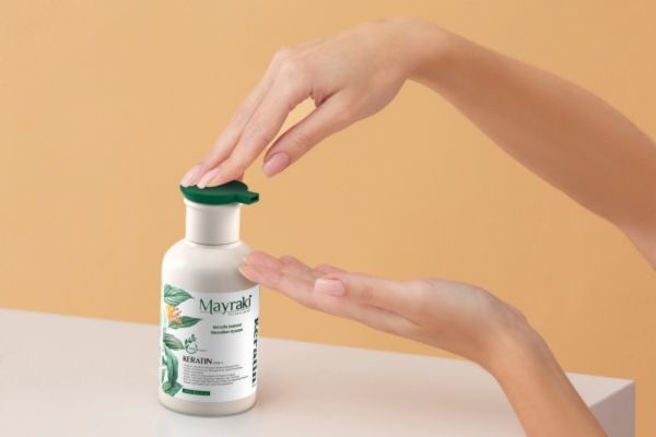 A person’s hands dispensing product from a white bottle of Mayraki Keratin Treatment with a green pump, featuring botanical illustrations on the label, against a soft beige background.