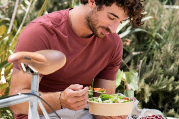 A curly-haired man in a red shirt enjoys a fresh salad while sitting outdoors next to a bicycle, promoting healthy eating habits as part of a balanced lifestyle.