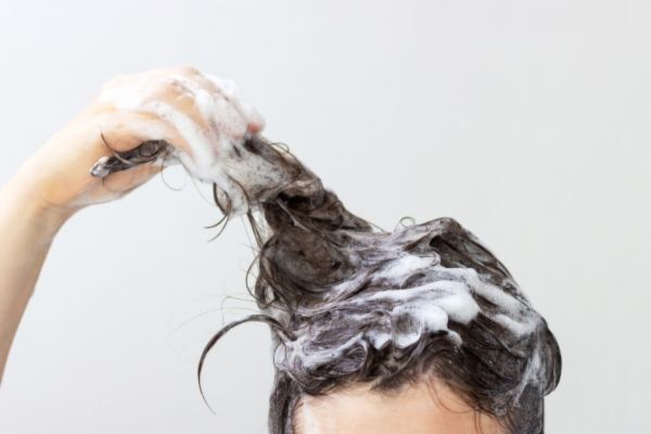 Close-up of a person washing their hair, with fingers massaging shampoo foam into wet dark hair against a plain light background.