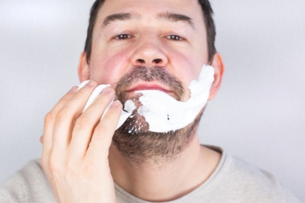 Close-up of a man with facial hair applying shaving cream to his beard area, preparing for grooming.