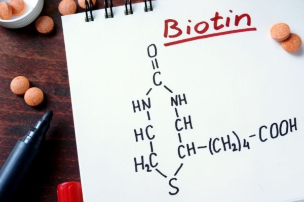 Notebook page with a hand-drawn chemical structure of biotin, labeled in red at the top, surrounded by round orange supplement tablets and a black marker on a wooden table.
