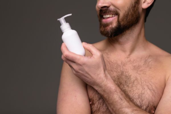 A shirtless man with a trimmed beard smiles while holding a white pump bottle, suggesting focus on key ingredients in beard leave-in conditioner or grooming products.