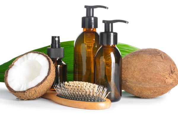 A natural hair care setup featuring amber-colored pump and spray bottles, a wooden hairbrush with light bristles, a whole coconut, a halved coconut showing its white flesh, and a green tropical leaf in the background. The scene emphasizes coconut-based or organic hair products with a clean, eco-friendly vibe.