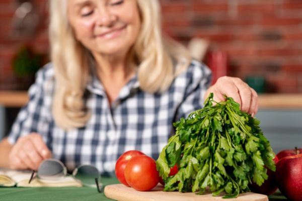 Lifestyle and Diet Tips to Slow Gray Hair An older woman with long gray-blonde hair smiles softly while seated at a kitchen table, gently holding a bunch of fresh parsley on a wooden cutting board. Surrounding the parsley are vibrant red tomatoes and apples, emphasizing a healthy diet tied to natural ways of slowing gray hair.
