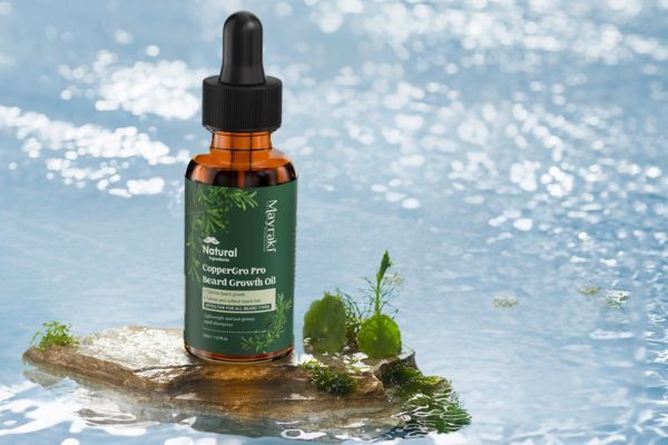 A bottle of Mayraki CopperGro Pro Beard Growth Oil stands on a small, mossy rock in the middle of shimmering water. The natural setting highlights the product's clean, botanical ingredients and eco-conscious grooming appeal.