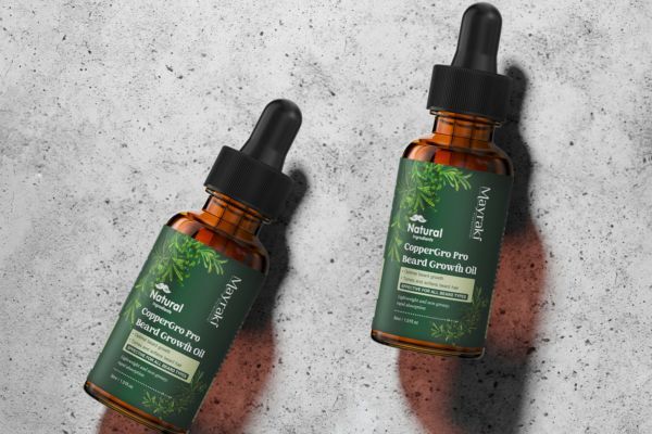 Two amber dropper bottles of Mayraki CopperGro Pro Beard Growth Oil lie on a textured concrete surface. The green-labeled packaging highlights that the product is all-natural and designed to promote beard growth with organic ingredients.