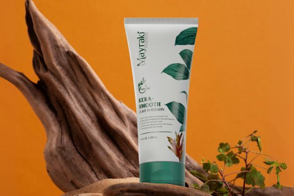 A tube of Mayraki Kera-Smooth Leave-in Keratin Hair Lotion is displayed against a warm orange background with natural wood and greenery. The botanical-themed packaging emphasizes its keratin-infused, plant-based formula for smoothing and strengthening hair.