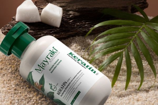 A bottle of Mayraki Professional Keratin Treatment resting on sand, surrounded by a palm leaf, wooden piece, and coconut chunks, evoking a tropical and natural vibe. The white and green packaging emphasizes botanical ingredients and a salon-quality hair care formula.