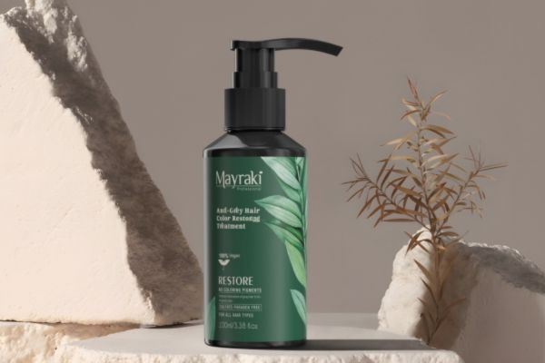 A bottle of Mayraki Anti-Grey Hair Color Restoring Treatment with a black pump and green label featuring leafy graphics, placed on a minimalist stone setup with natural decor. The product claims to be 100% vegan and free of parabens, sulfates, and silicones.