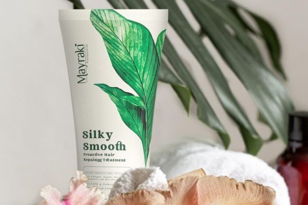 A tube of Mayraki Silky Smooth Proactive Hair Repairing Treatment stands upright amid soft towels, pink flowers, and a leafy backdrop. The packaging features a bold green leaf design, highlighting its natural ingredients and focus on repairing damaged hair.