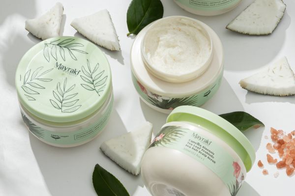 Open and closed jars of Mayraki hair or scalp treatment are arranged with coconut chunks, green leaves, and pink Himalayan salt on a white surface. The pastel green packaging and natural elements emphasize the product's botanical, nourishing ingredients for scalp health.