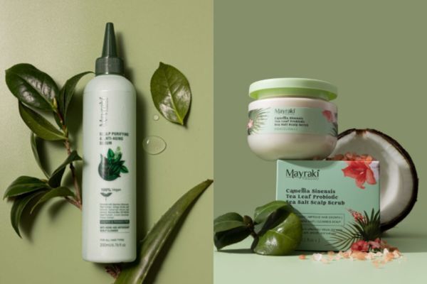 Hair Mayraki products, including a scalp purifying serum and Camellia Sinensis Tea Leaf Probiotic Sea Salt Scalp Scrub, are displayed against a green background with fresh leaves and a halved coconut. The setup emphasizes Hair Mayraki’s plant-powered approach to scalp health and cleansing.