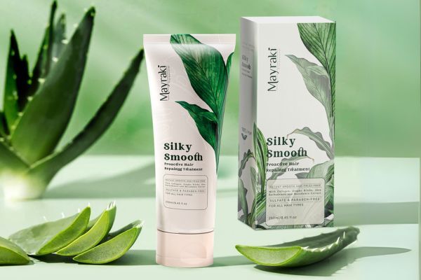 A tube and box of Mayraki Silky Smooth Proactive Hair Repairing Treatment are displayed on a light green surface with aloe vera leaves around them. The packaging features botanical artwork and emphasizes that the product is sulfate- and paraben-free, suitable for all hair types.