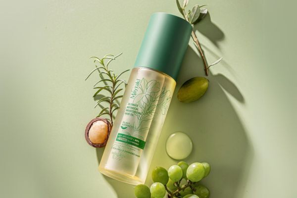 A bottle of Mayraki Ultimate Botanical Moisturizing Hair Oil lies on a soft green surface surrounded by natural ingredients like grapes, olives, rosemary, and macadamia. The elegant packaging and botanical elements highlight the product’s plant-based, hydrating formula for hair care.